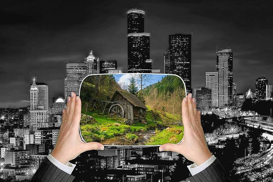 zwei Hände halten einen Bildschirm mit grüner Landschaft, im Hintergrund eine graue Großstadt