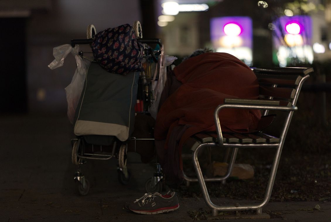 Person schläft unter einem Schlafsack auf einer öffentlichen Bank, davor ein Einkaufswagen mit Habseeligkeiten